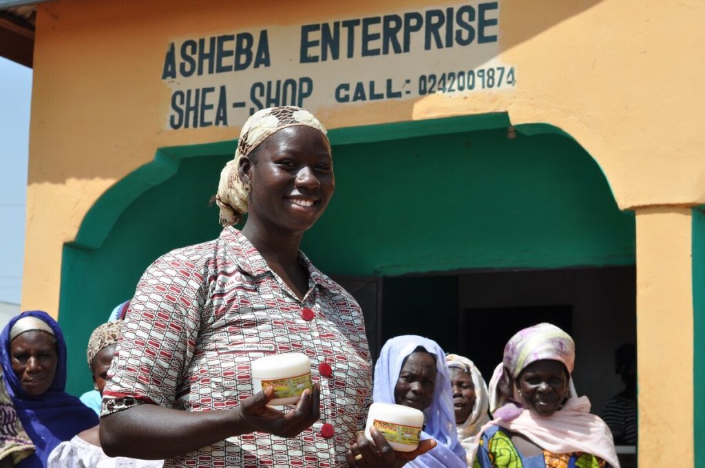 A Ghanaian woman smiles and stands outside her shea butter shop.