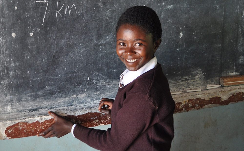 Rockcilia is a student in Zambia, she stands at the chalkboard in a classroom