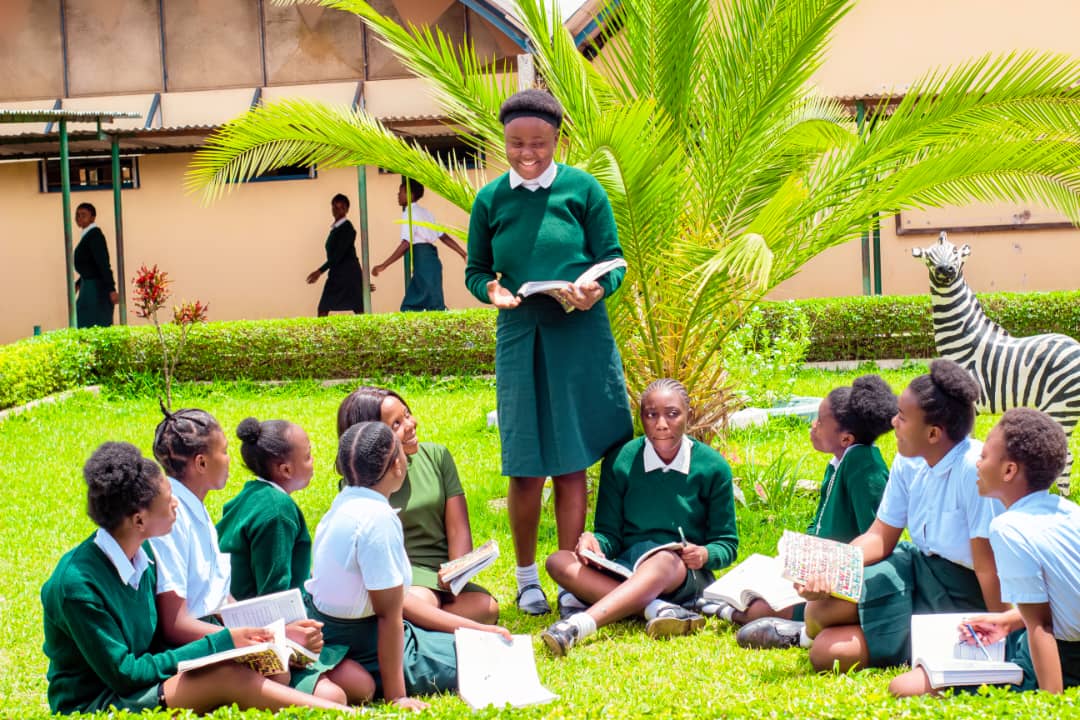 Standing student Theresa, in uniform, holds a book and presents to seated Zambian woman Harriet and a group of uniformed students on the grass outdoors in Zambia.