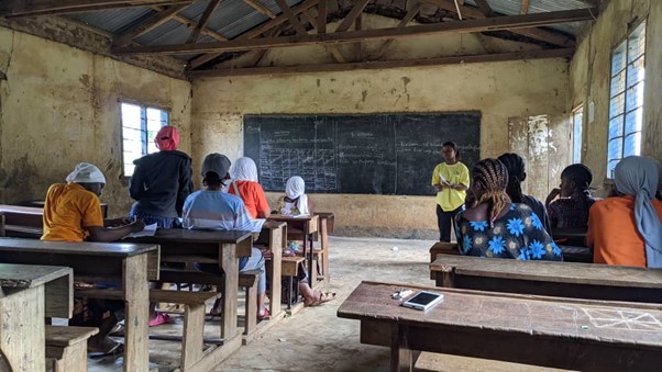 Sara, a Tanzanian woman in a yellow top and black trousers standing in front of a chalk board leading a Business Guide session for community members.