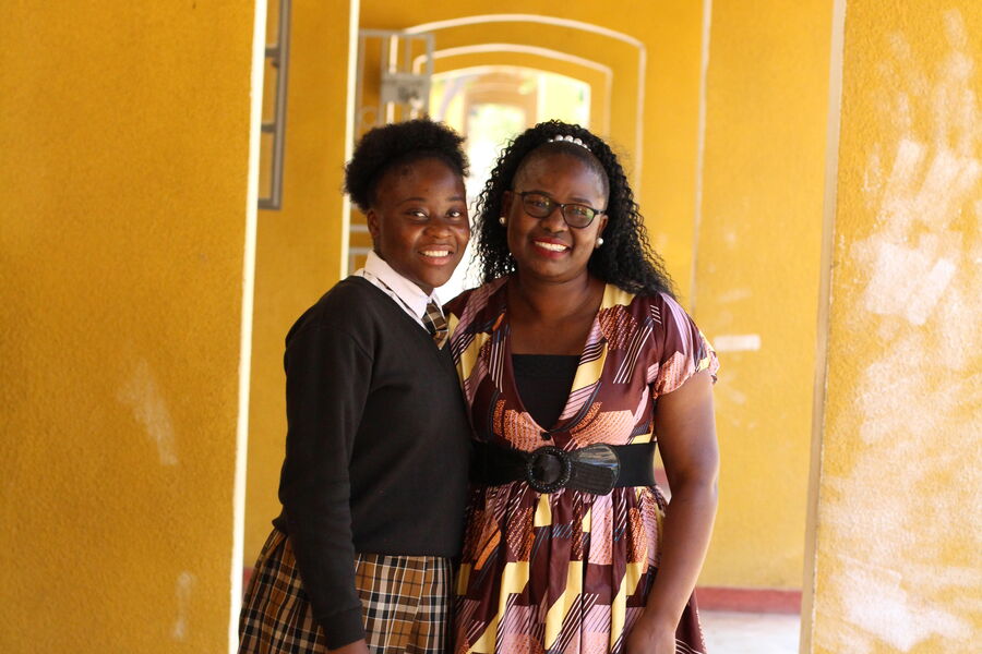 Mapalo, a female Zambian student stands with her Teacher Mentor, Susan and smiles, as they stand in a yellow school corridor.