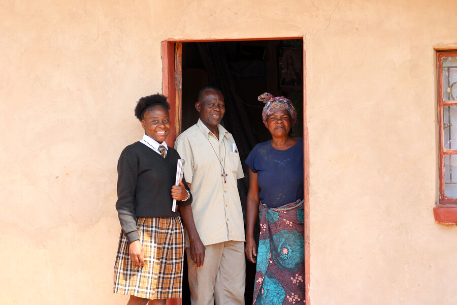 Mapalo, a Zambian student wearing school uniform and holding a book, stands with her father and step-mother outside their home in Mpika District, Zambia.