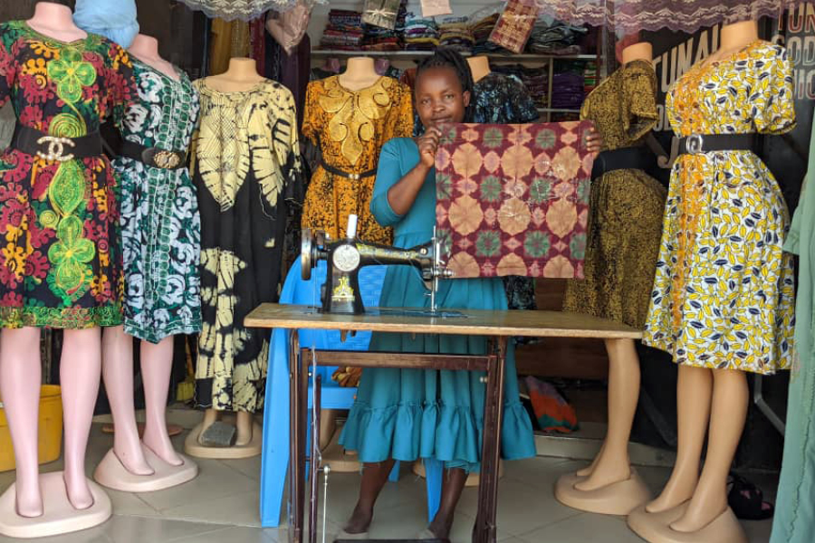 Anita, a Tanzanian woman stands in her clothing shop behind a sewing machine, and holds up colorful fabric.