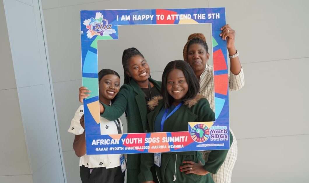 Four smiling Zambian women pose with a photo frame at the African Youth Sustainable Development Goals (SDG) Summit in Lusaka, Zambia.