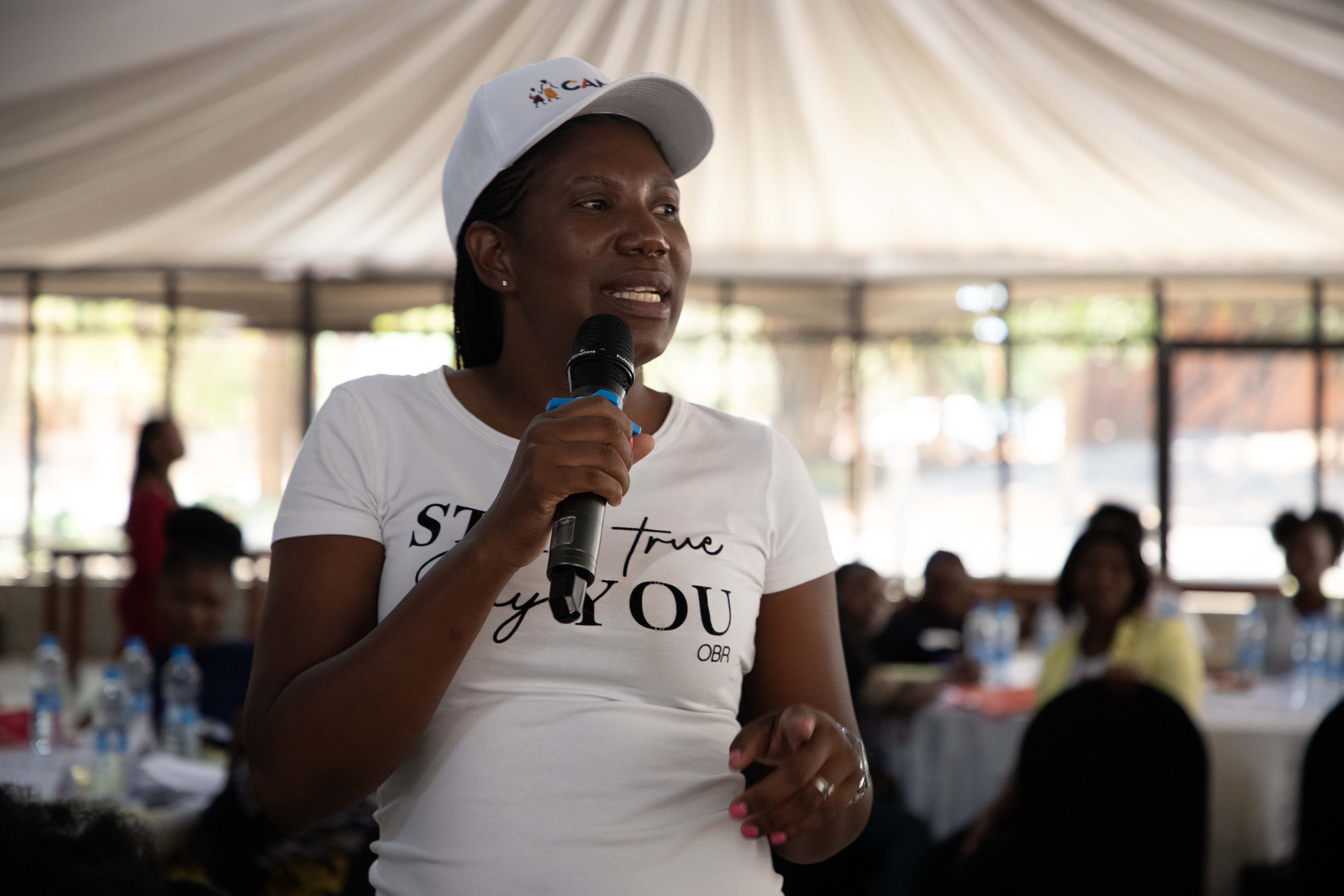 Mwangala, a Zambian woman wearing a white t shirt and cap speaking into a microphone.